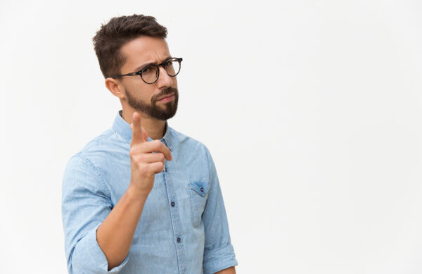 Frowning concerned guy shaking finger at camera. Handsome young man in casual shirt and glasses standing isolated over white background. Warning gesture concept