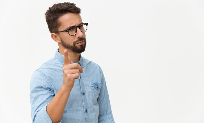 Frowning concerned guy shaking finger at camera. Handsome young man in casual shirt and glasses standing isolated over white background. Warning gesture concept