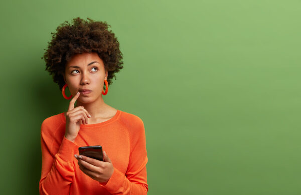 Photo of pretty ethnic woman ponders on how to answer question, thinks deeply about something, uses modern mobile phone, tries to made up good message, keeps index finger near lips, stands indoor