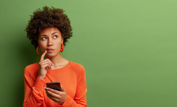 Photo of pretty ethnic woman ponders on how to answer question, thinks deeply about something, uses modern mobile phone, tries to made up good message, keeps index finger near lips, stands indoor